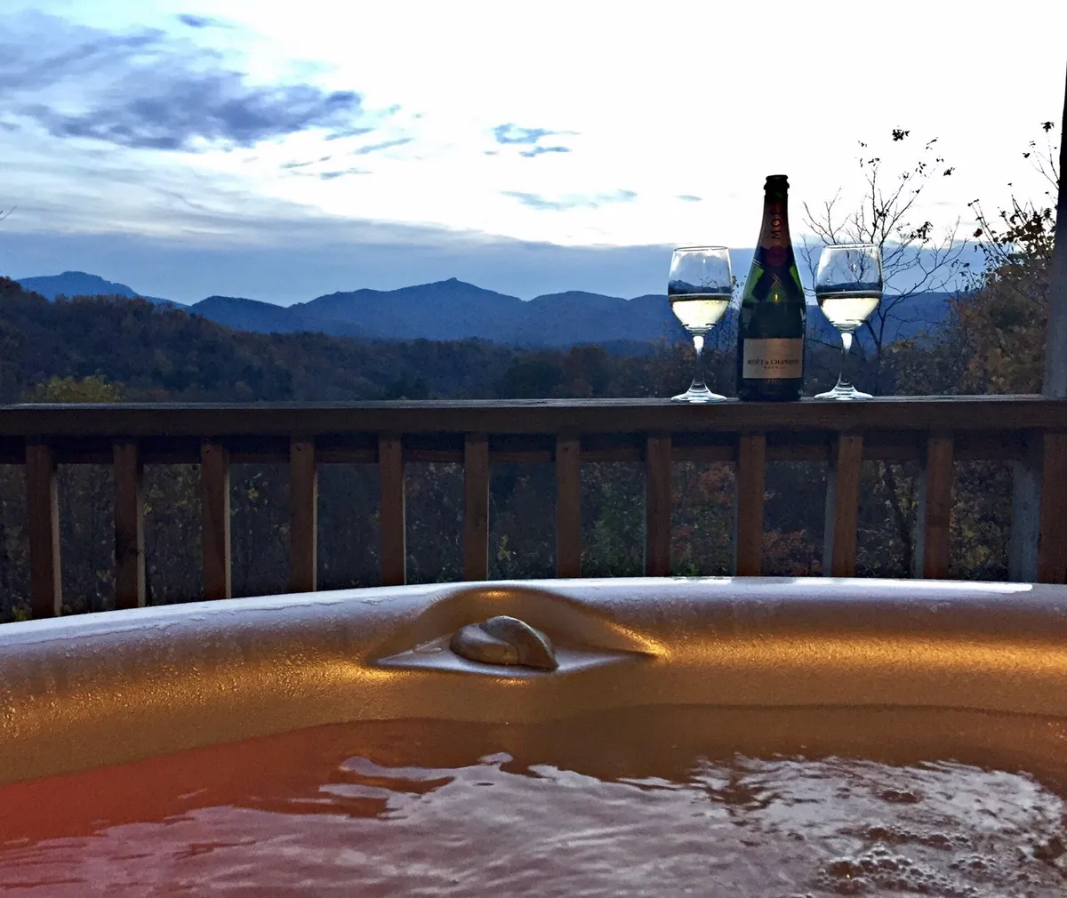 The photo features a private hot tub on the deck, champagne glasses poised on the edge, and a sweeping view of the mountains beyond. The mood is intimate and serene, perfect for relaxation or group celebration as the sun sets.