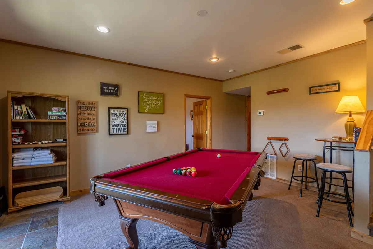 This image features a pool table as the central focus, surrounded by a bookshelf, bar stools, and a table lamp. The scene evokes a relaxed atmosphere perfect for friendly games and socializing, while the nearby bookshelf hints at activities beyond billiards.