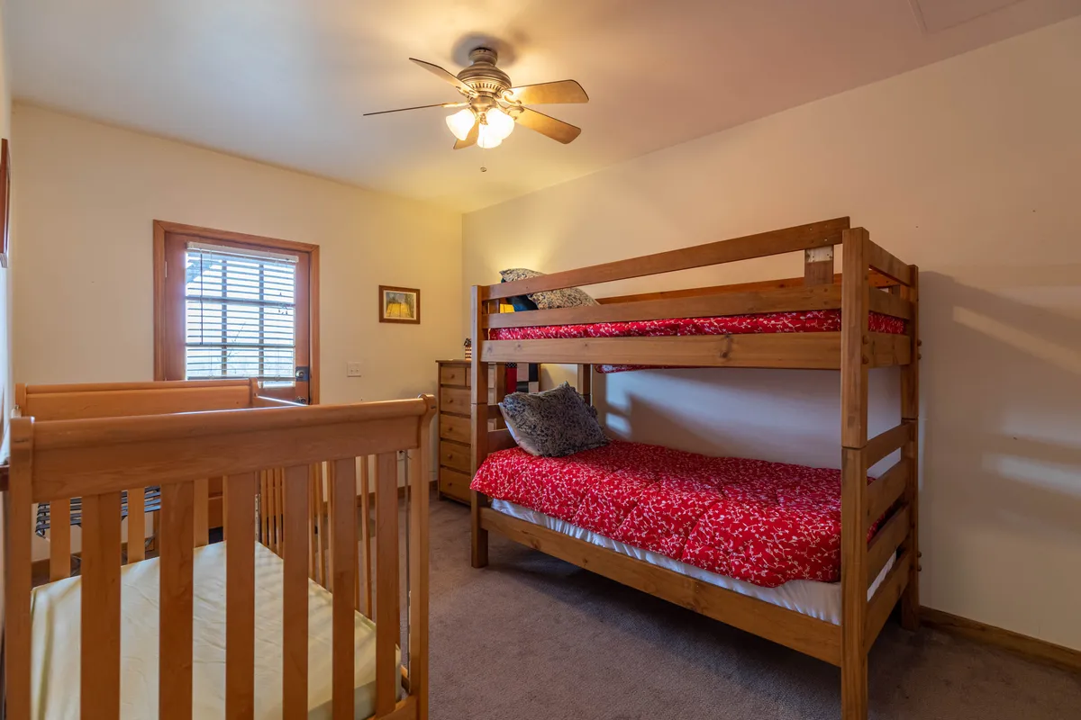 A cozy bedroom featuring bunk beds, a crib tucked nearby, a ceiling fan overhead, and a window with blinds. The room has warm lighting and family-friendly decor, ideal for accommodating children and flexible sleeping arrangements.