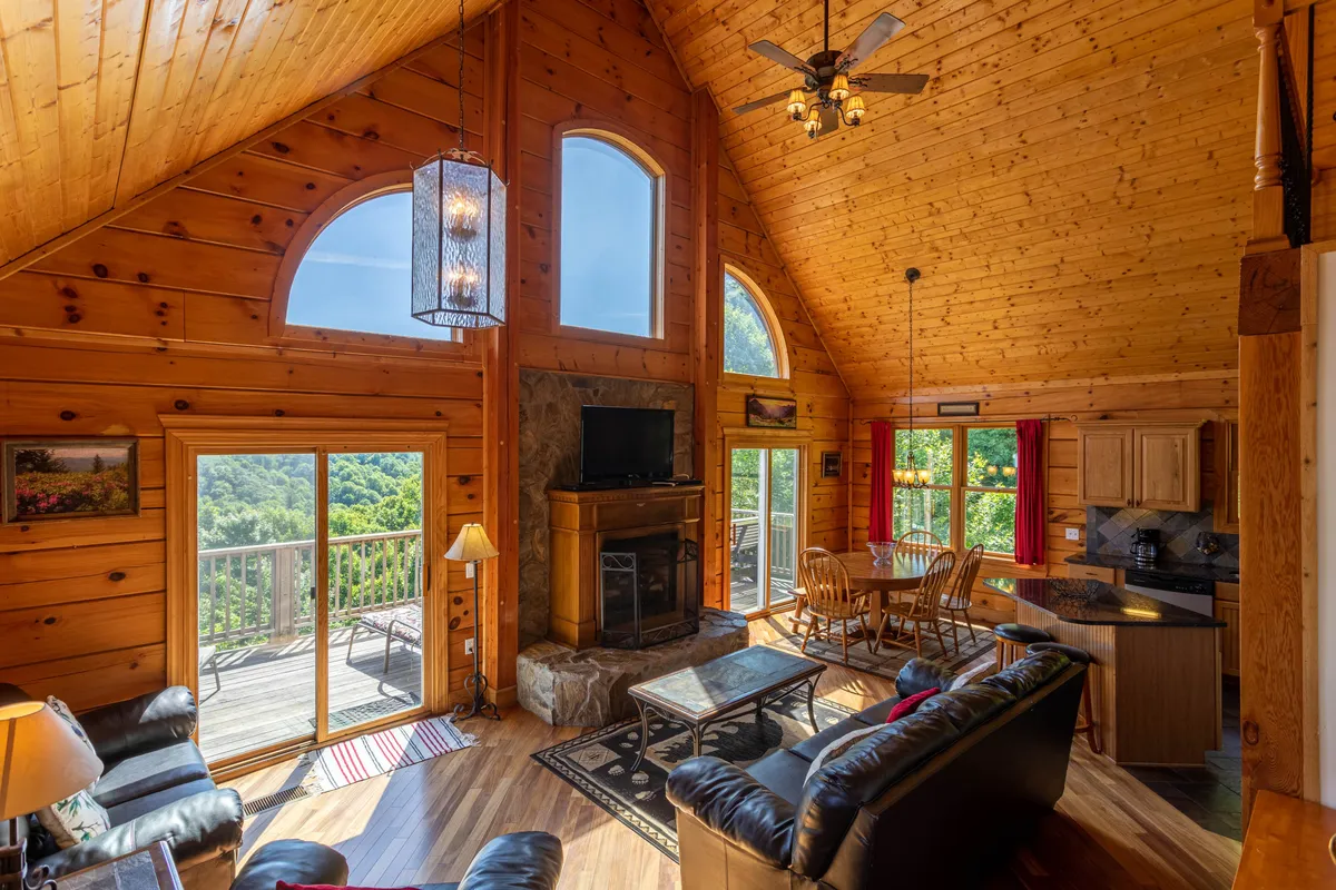 A spacious living and dining area characterized by large windows offering scenic mountain views. The stone fireplace and exposed wooden beams create a cozy mountain ambiance, while the wooden dining table encourages shared meals and connection. This open-plan space bridges relaxation and communal dining, seamlessly incorporating the outdoors through its windows.