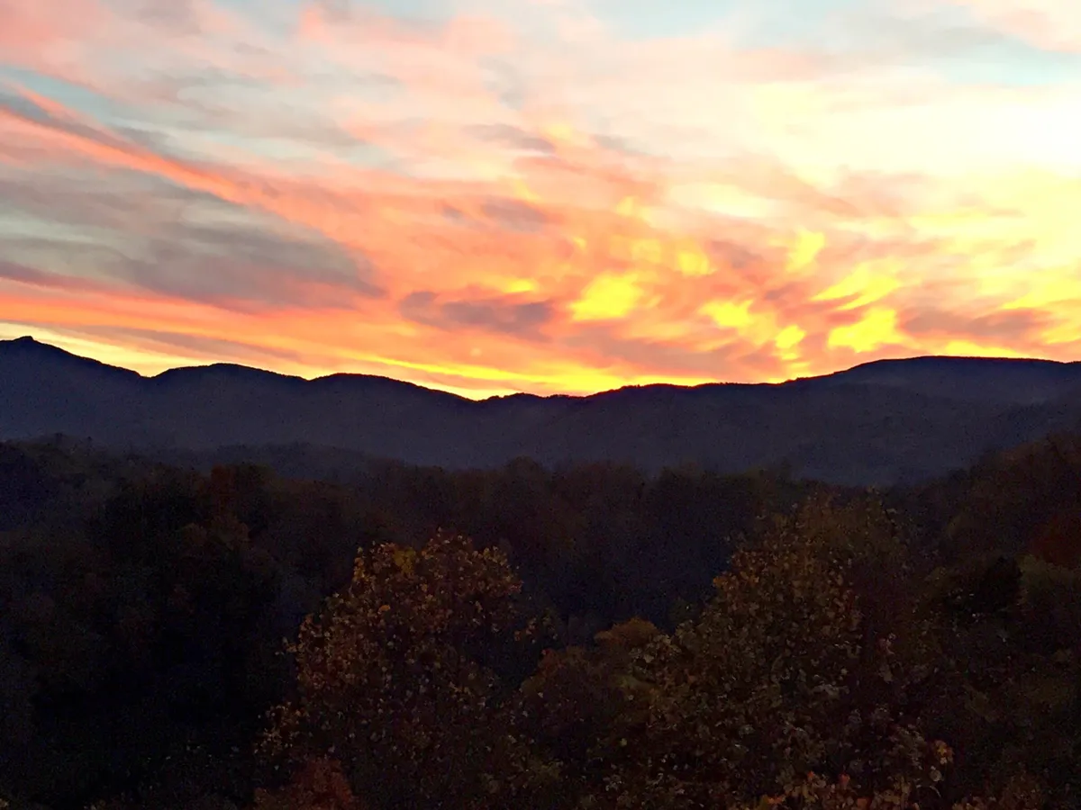 A stunning view of a mountain silhouette against a vibrant sunset sky, with colorful clouds and a dense tree line. The mood is serene and romantic, inviting guests to appreciate the natural beauty and peacefulness of the Blue Ridge Mountains.
