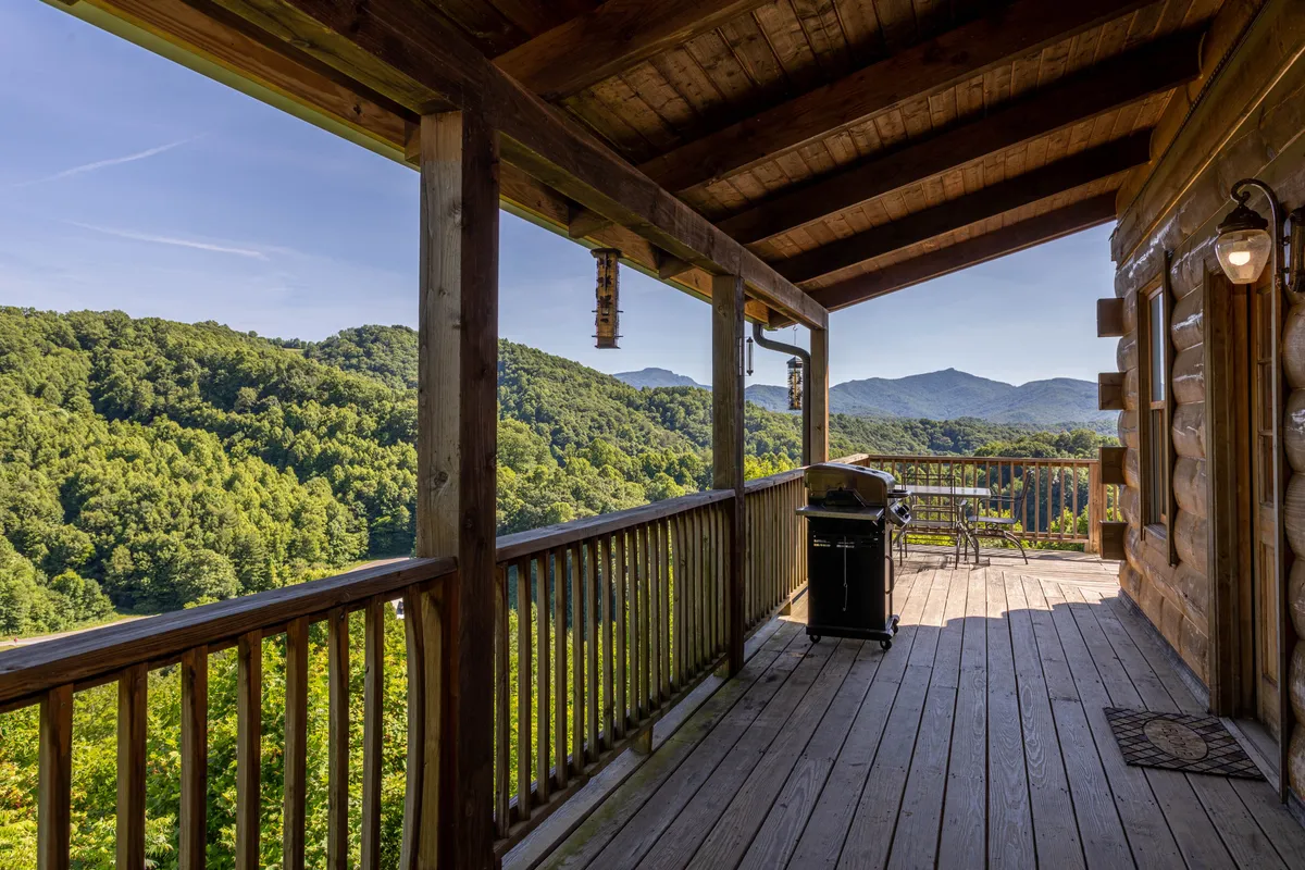 This image displays a wooden deck with outdoor seating and a gas grill, set against a stunning mountain view. The scene is tranquil, with soft lighting suggesting either morning or evening, and the deck is clearly designed for relaxation and al fresco dining.