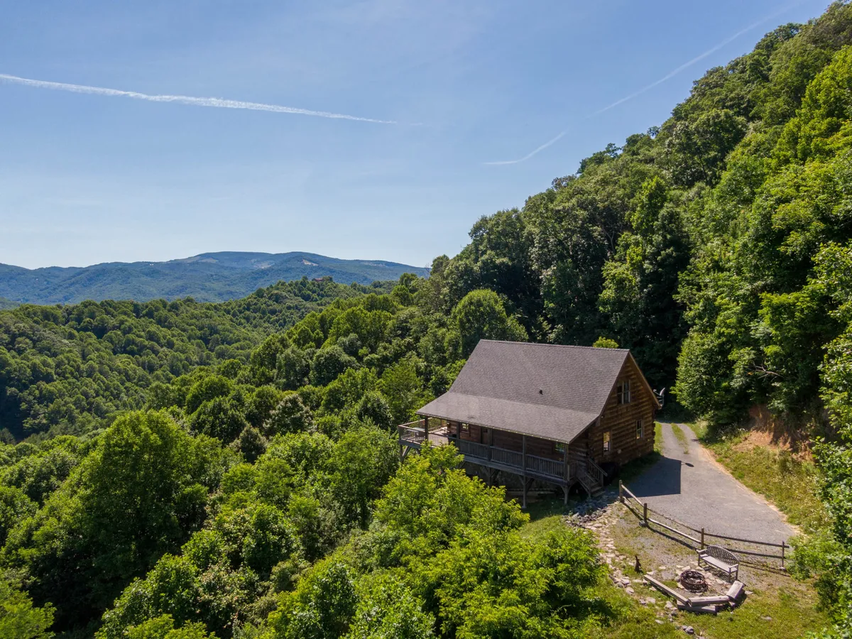 This image presents the log cabin nestled among tall trees with a clear view of the surrounding Blue Ridge Mountains. A welcoming fire pit area and a spacious deck hint at outdoor family gatherings and tranquil evenings under the sky.
