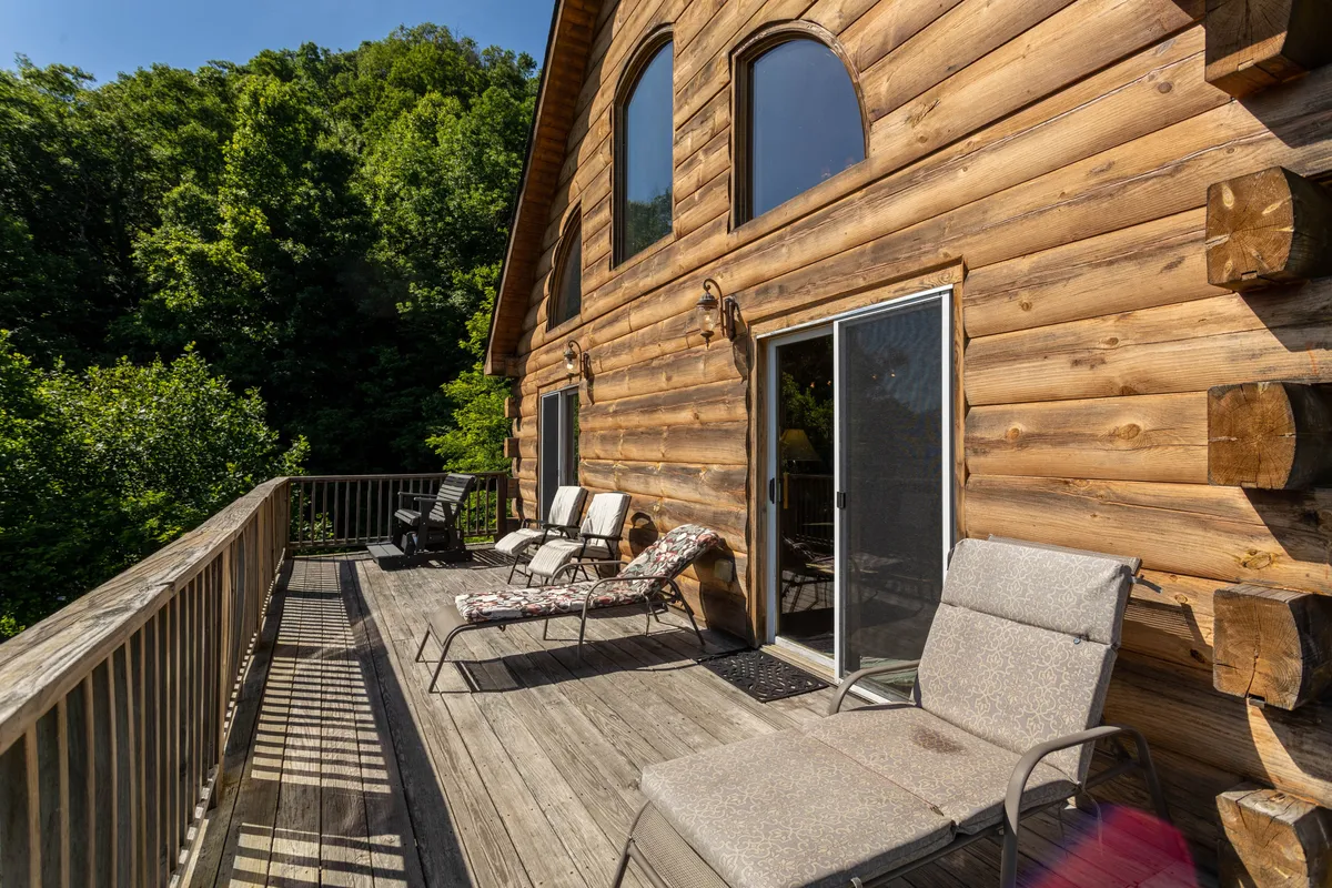 A log cabin exterior with a spacious wooden deck, lounge chairs for relaxing, and lush greenery surrounding the area. The deck invites guests and pets alike to relax, enjoy nature, and take in the sunset.