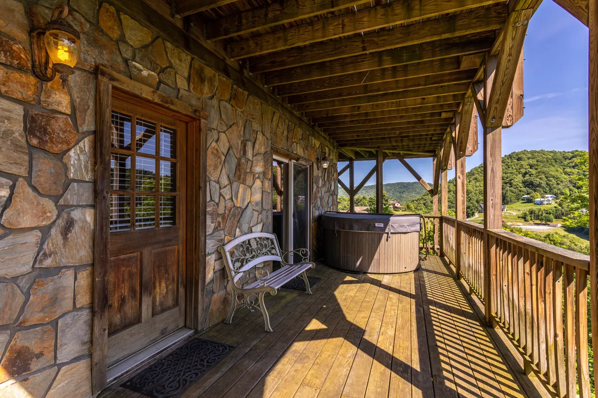 An exterior lower deck scene with a stone wall, bubbling hot tub, wooden bench, and a broad view of the mountains. The area is designed for relaxation and socializing, seamlessly connecting the interior entertainment spaces to the fresh mountain air outdoors.