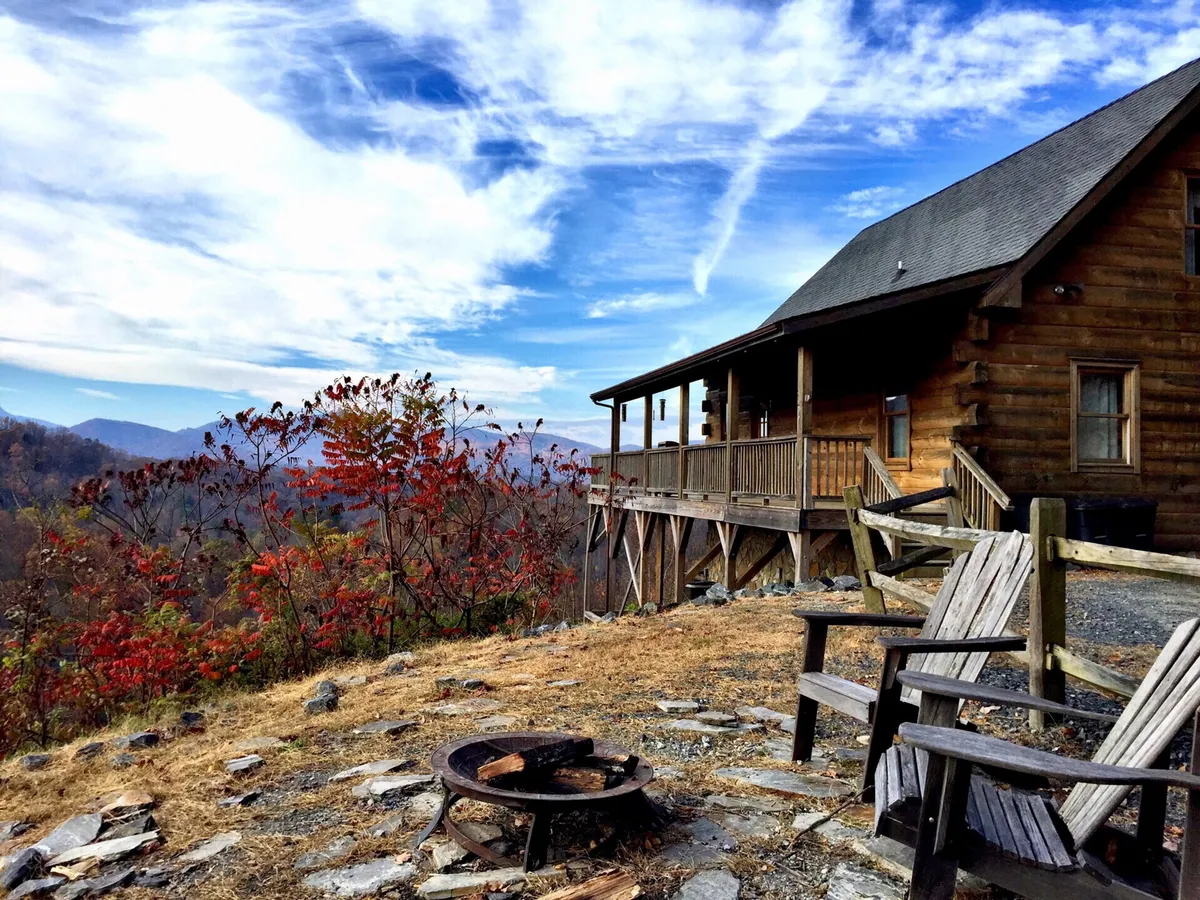The log cabin is pictured amid vibrant autumn foliage, with a cozy fire pit and wooden chairs facing panoramic mountain views. The setting radiates warmth and invites families to unwind together outdoors.