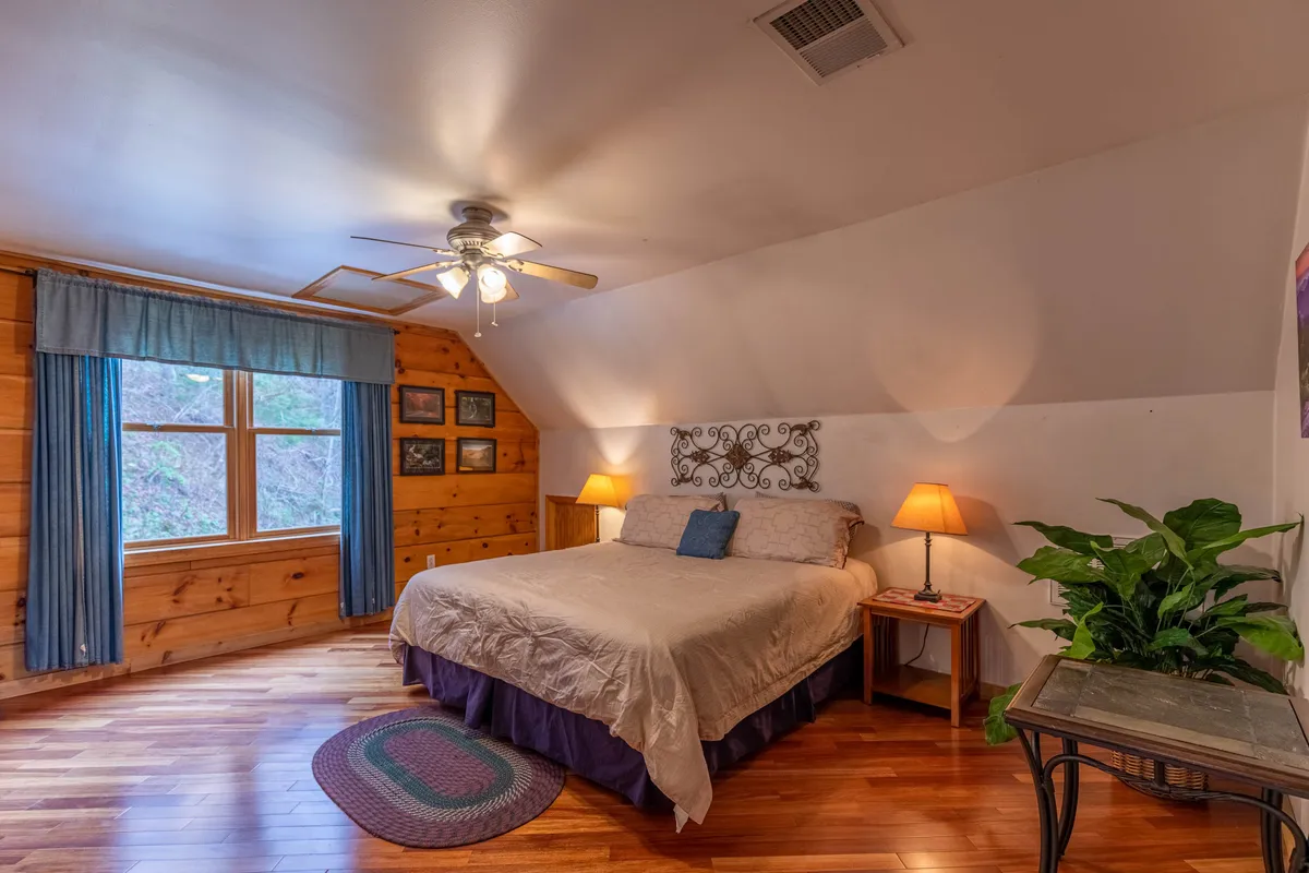 A spacious bedroom featuring a decorative headboard, a large window with curtains allowing in natural light, nightstands with lamps, and a touch of greenery from a potted plant. The decor is cozy and welcoming.
