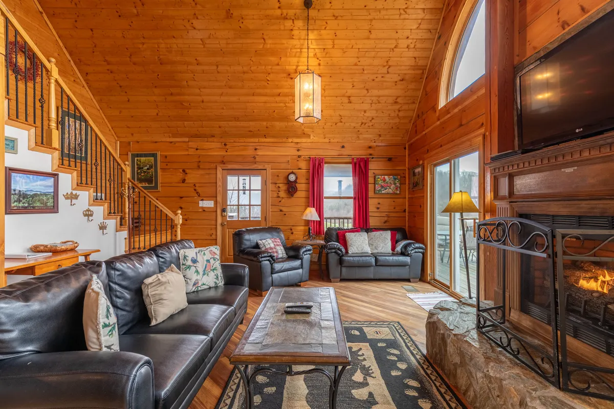 A sunlit living room with soaring cathedral ceilings, rustic wooden beams, and expansive windows that frame panoramic mountain views. The stone gas-log fireplace is the heart of the room, surrounded by plush leather sofas, creating a cozy gathering spot for families or friends. The room exudes warmth and comfort, inviting both relaxation and lively conversation.