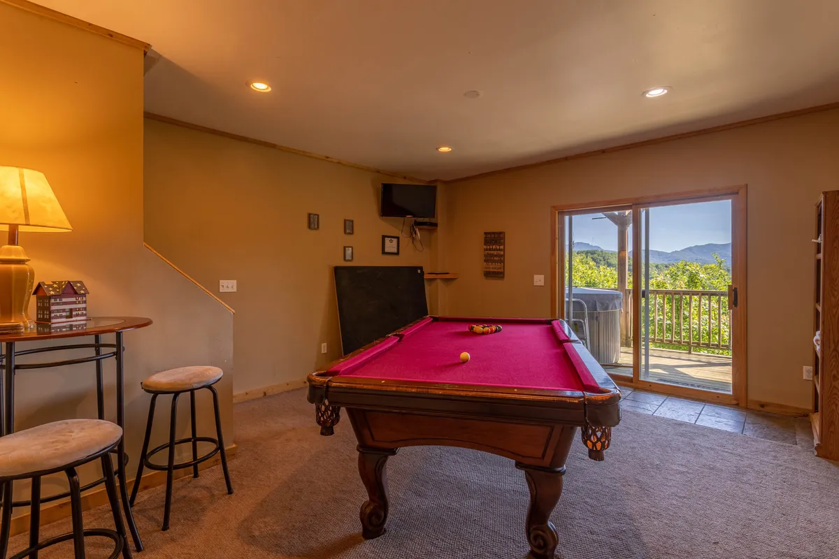 A lively interior scene centered on a pool table, with a sliding glass door hinting at mountain views outside. Bar stools and a lamp create a casual, fun ambiance for friendly competition and group games after outdoor adventures.